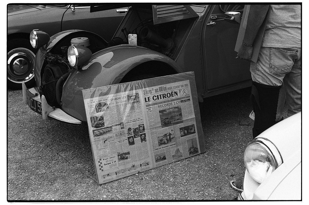 Une voiture ancienne Citroën 2CV garée, avec une affiche de journal posée devant, montrant des articles et des images sur la voiture.