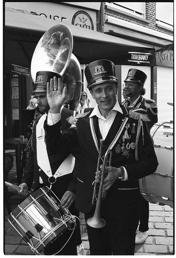 Un groupe de musiciens lors d'un défilé, l'un d'eux, en uniforme, salue la caméra tout en tenant une trompette.