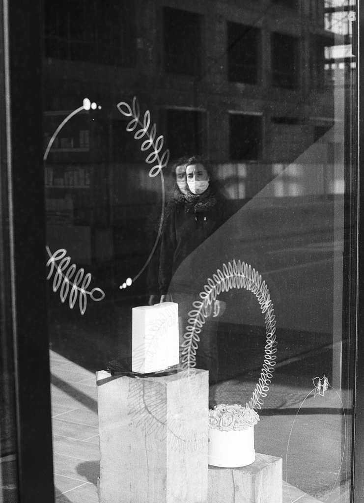 Reflet d'une personne portant un masque dans une vitrine avec des éléments décoratifs, photographié en noir et blanc.