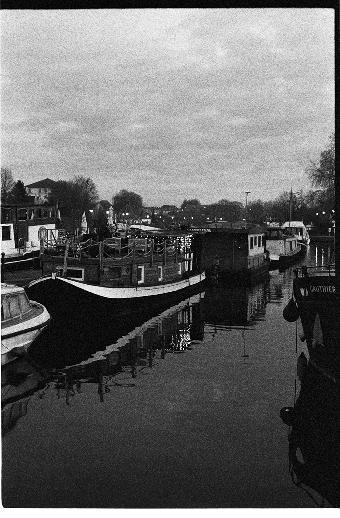 Photographie en noir et blanc de bateaux amarrés le long d'un canal, reflétés dans l'eau calme sous un ciel nuageux.