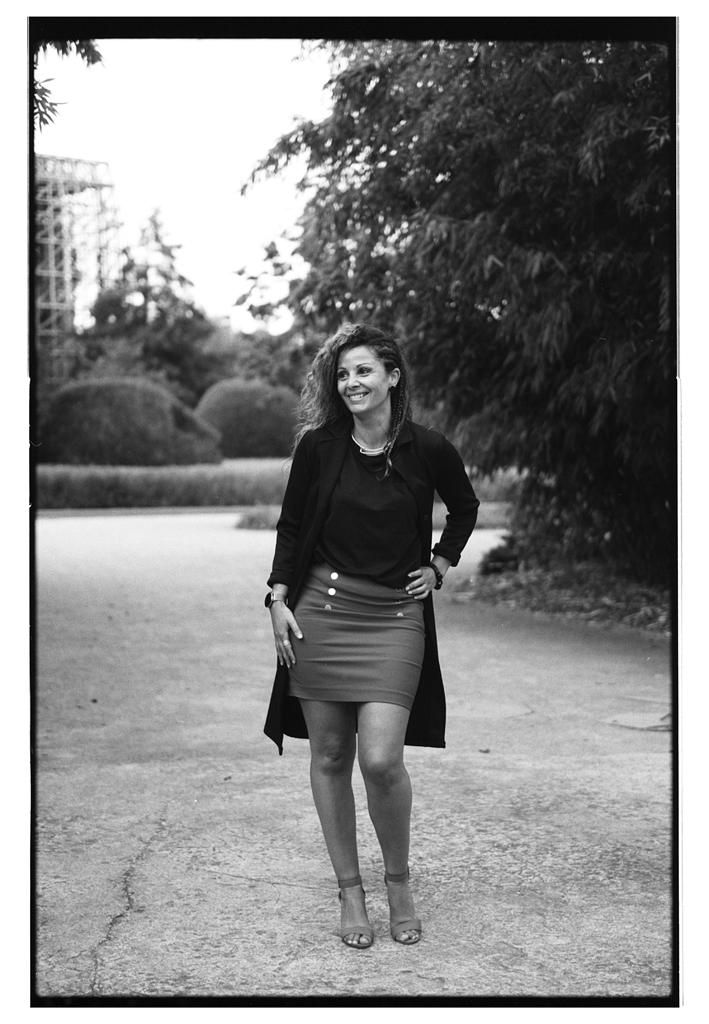 Portrait d'une femme souriante, debout sur un chemin, portant une jupe courte et un haut noir, avec des arbres en arrière-plan.