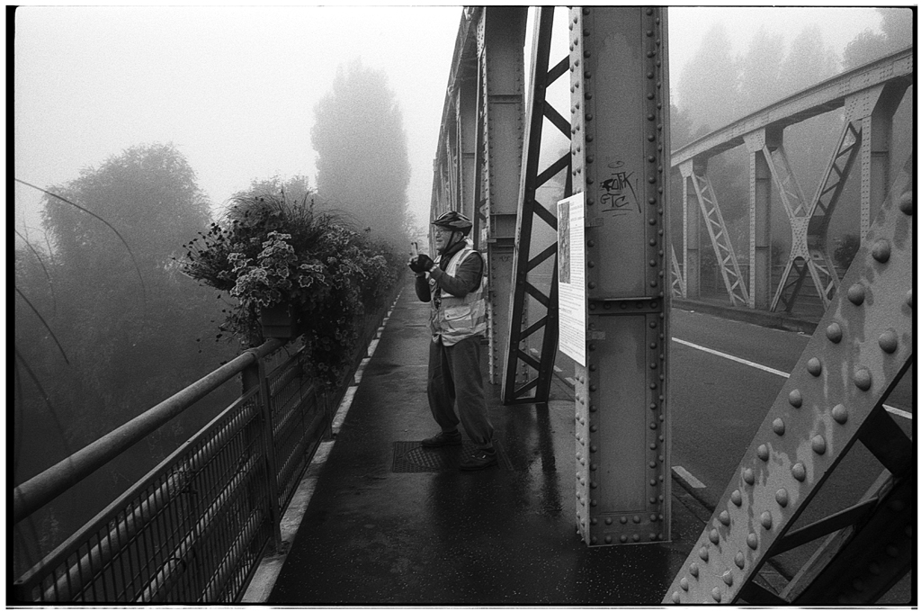 Un photographe en train de prendre une photo sur un pont en métal, sous un ciel brumeux, avec des fleurs sur le garde-corps.