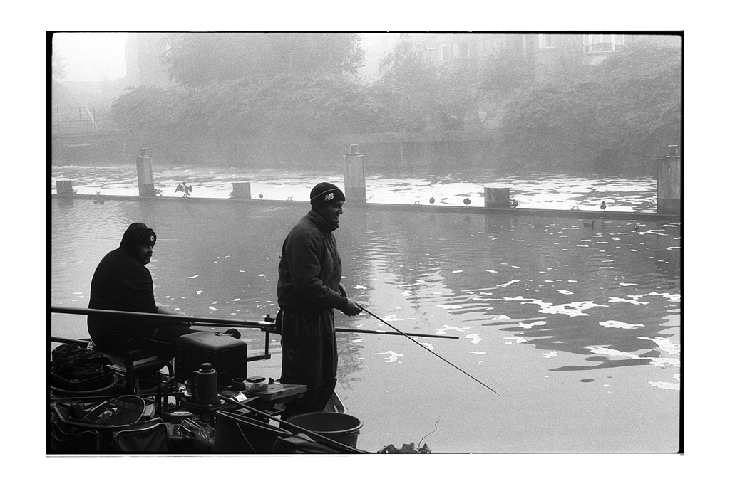 Deux pêcheurs silhouettes au bord d'une rivière brumeuse, photographiés en noir et blanc.