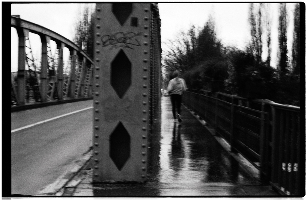 Une personne marchant sur un chemin humide près d'un pont en métal, sous un ciel nuageux, capturée en noir et blanc.