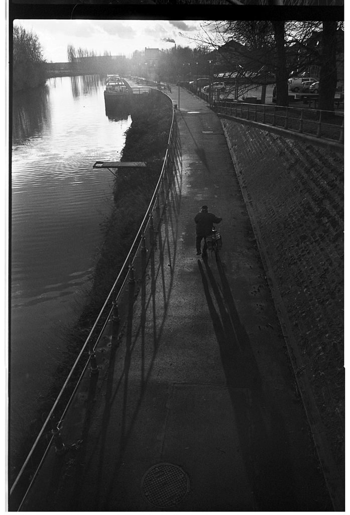 Un homme poussant un vélo le long d'une promenade au bord d'une rivière, avec des ombres longues projetées sur le sol.