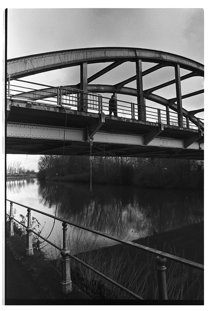 Vue en noir et blanc d'un homme marchant sur un pont métallique surplombant une rivière, avec des arbres en arrière-plan.