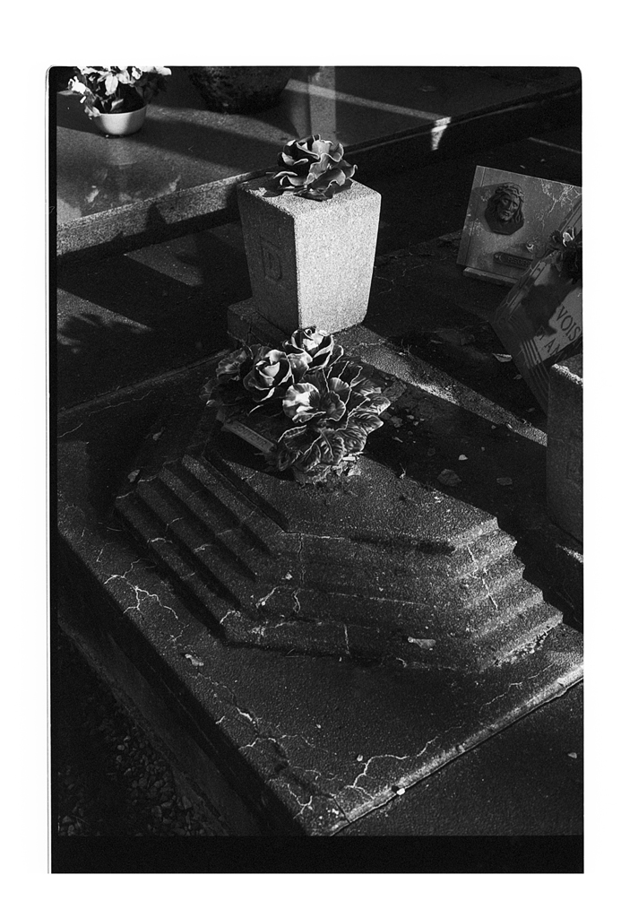 Une pierre tombale sculptée, recouverte de fleurs, dans un cimetière, photographiée en noir et blanc avec des jeux d'ombres et de lumière.