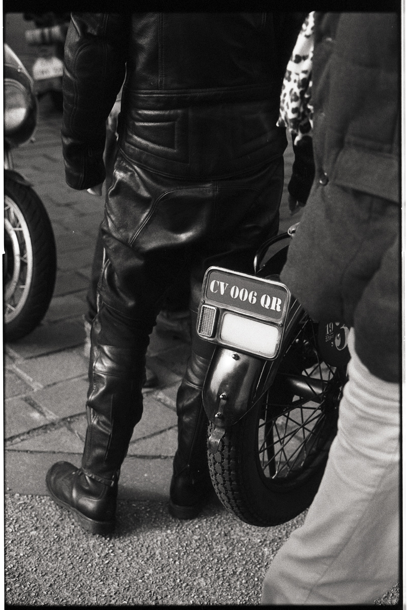 Un motard en tenue en cuir observant une moto, avec un groupe de personnes en arrière-plan. Photo argentique au 28mm.