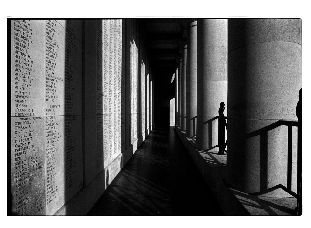 Vue en noir et blanc d'un couloir avec des colonnes, entouré de murs portant des inscriptions. Des ombres distinctes se projettent sur le sol, créant un contraste saisissant entre la lumière et l'obscurité.