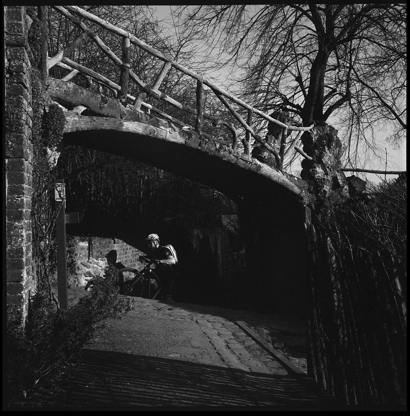 Un cycliste passant sous un pont en bois, entouré d'arbres dénudés et de feuillages, capturé en noir et blanc.