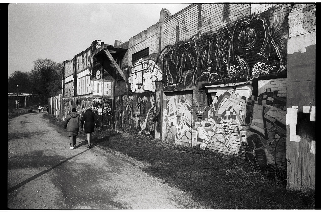Une rue déserte bordée de murs recouverts de graffitis, avec deux personnes marchant sur un chemin de terre. Photo au 28mm.
