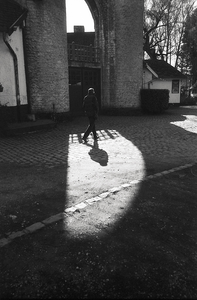 Une silhouette d'une personne marchant sur un chemin pavé, avec des ombres créées par une lumière traversant un arc en pierre.