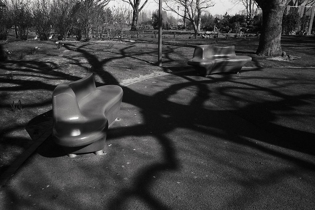 Deux bancs en plastique dans un parc, projetant des ombres sur le sol, avec des arbres en arrière-plan.