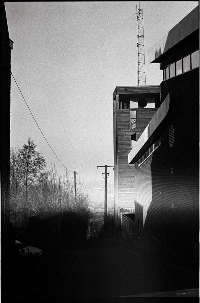 Une rue descendante avec des murs de bâtiments modernes, une antenne en hauteur, et des arbres en arrière-plan, capturée en noir et blanc.