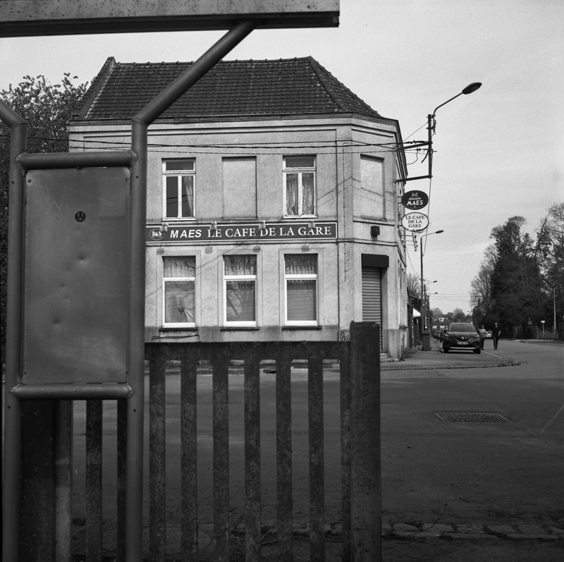 Vue en noir et blanc d'un café nommé 'Le Café de la Gare', avec des fenêtres visibles et un panneau éclairé, encadré par une clôture en bois.