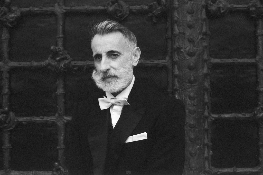 Portrait en noir et blanc d'un homme barbu, vêtu d'un costume élégant, avec un nœud papillon, posé devant une porte en métal ornée.