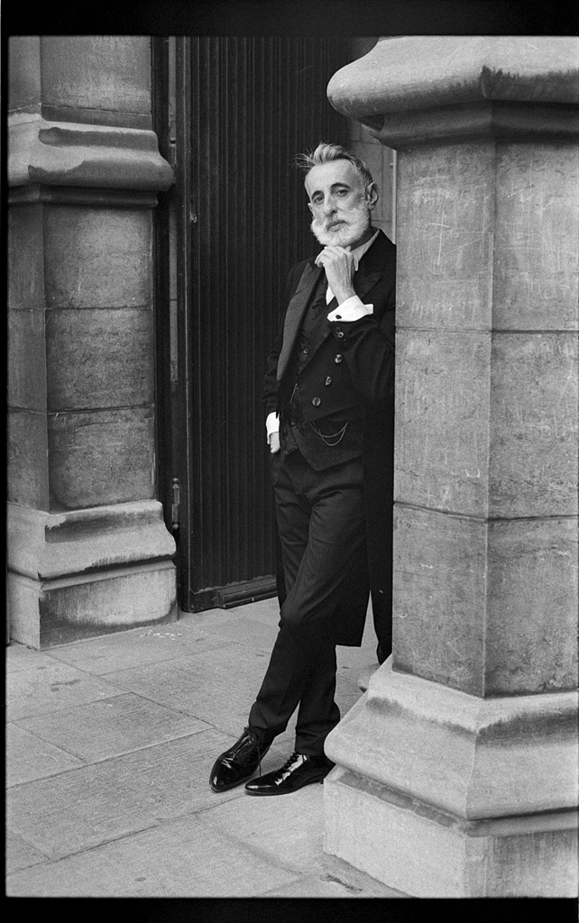 Portrait en noir et blanc d'un homme avec une barbe et des cheveux gris, vêtu d'un costume historique, posé contre une colonne dans une rue de Lille.