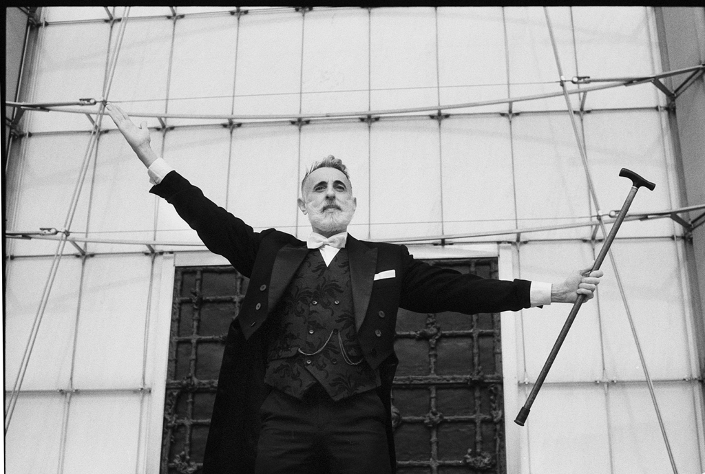 Portrait en noir et blanc d'un homme en costume d'époque, bras écartés, tenant une canne, devant un fond de bâtiment ancien.