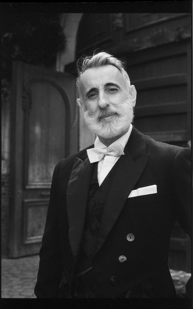 Portrait en noir et blanc d'un homme avec une barbe grise et des cheveux coiffés en arrière, vêtu d'un costume noir élégant avec un nœud papillon blanc, pris devant une porte ancienne à Lille.