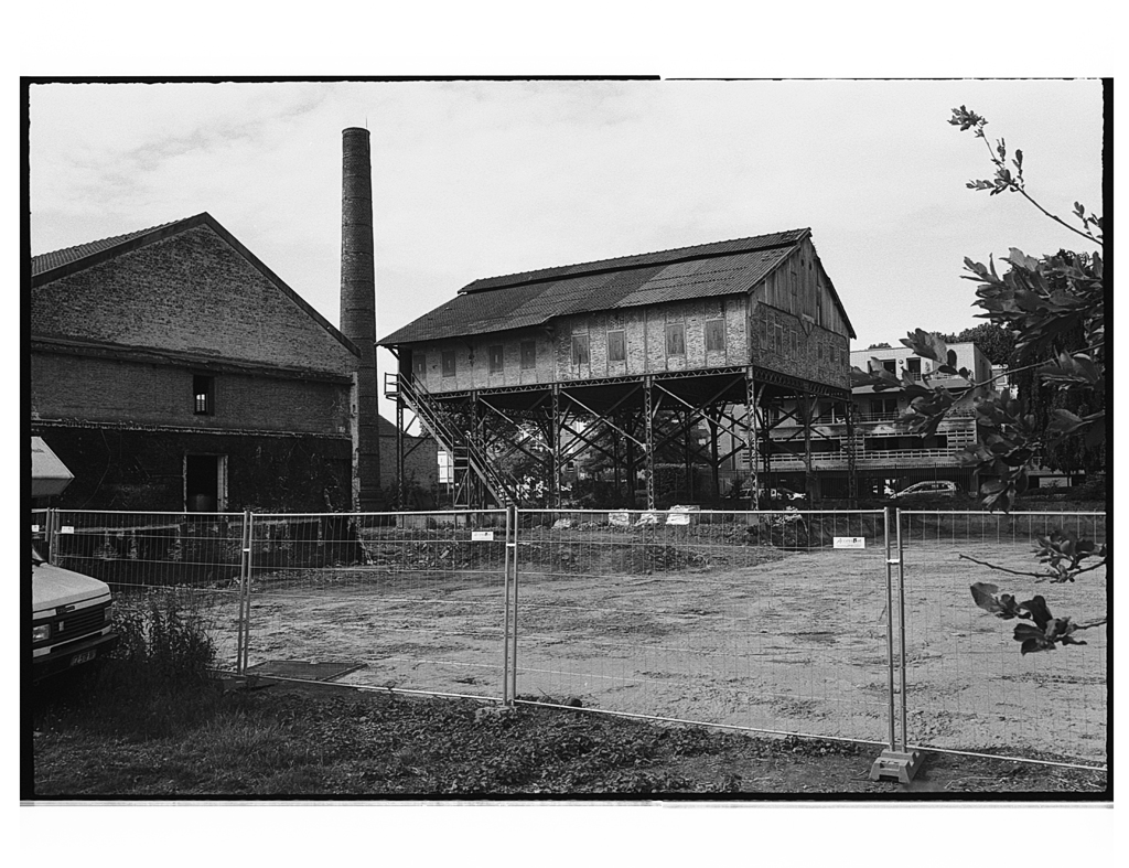 Une ancienne structure en bois sur pilotis, à côté d'un bâtiment en brique, avec une cheminée en arrière-plan. Des clôtures de chantier entourent le site, laissant entrevoir un terrain dégagé.