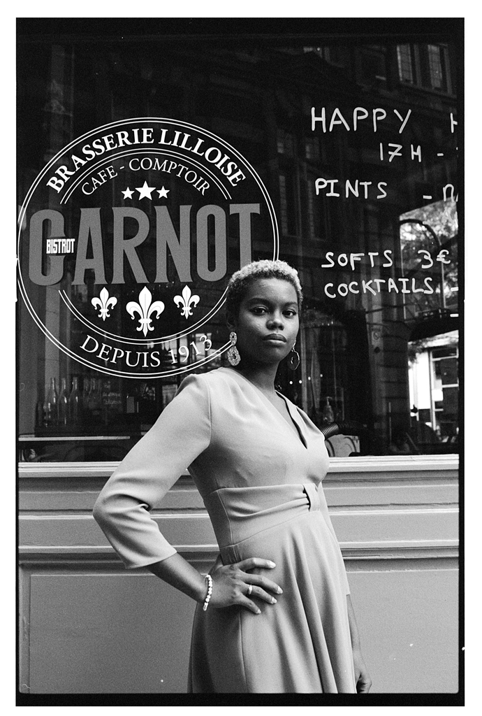 Portrait noir et blanc d'une femme debout devant une brasserie avec un enseigne visible en arrière-plan.