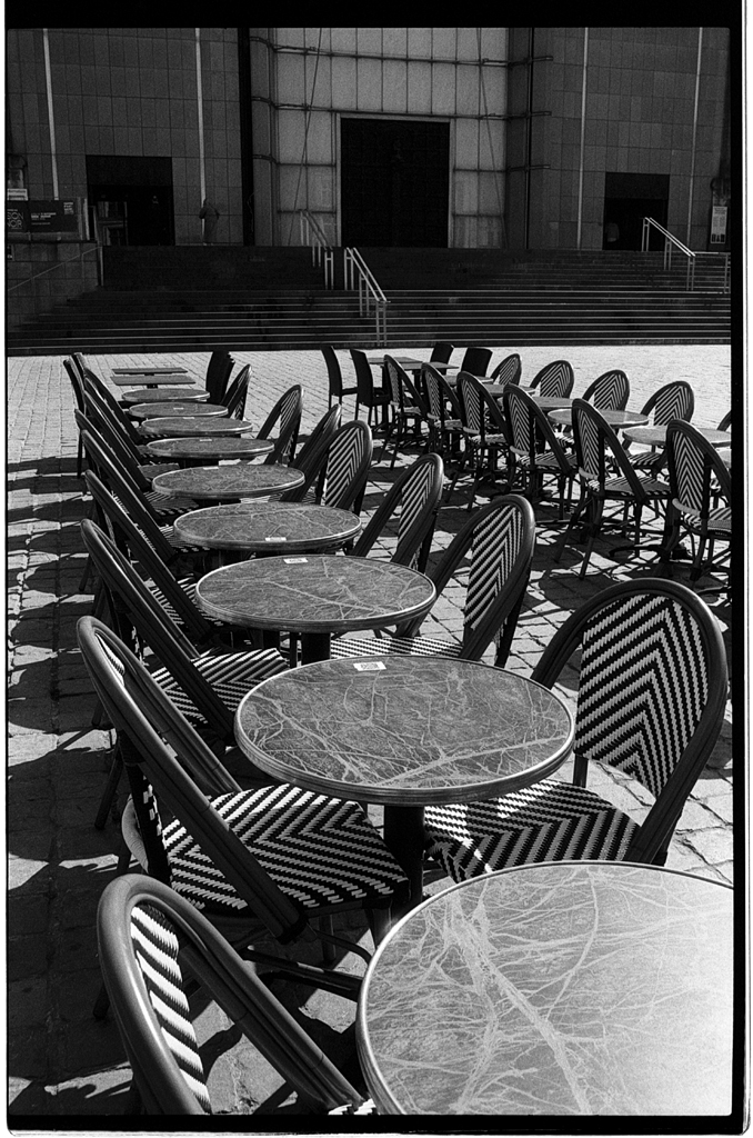 Des tables et chaises vides en noir et blanc, disposées en rangées sur une surface pavée, devant un bâtiment.
