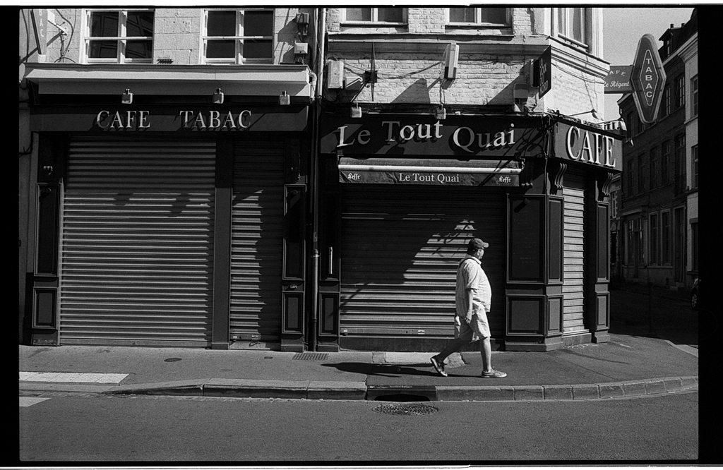 Un homme marche devant des cafés fermés, avec des volets roulant en métal sur des façades de bâtiments anciens.