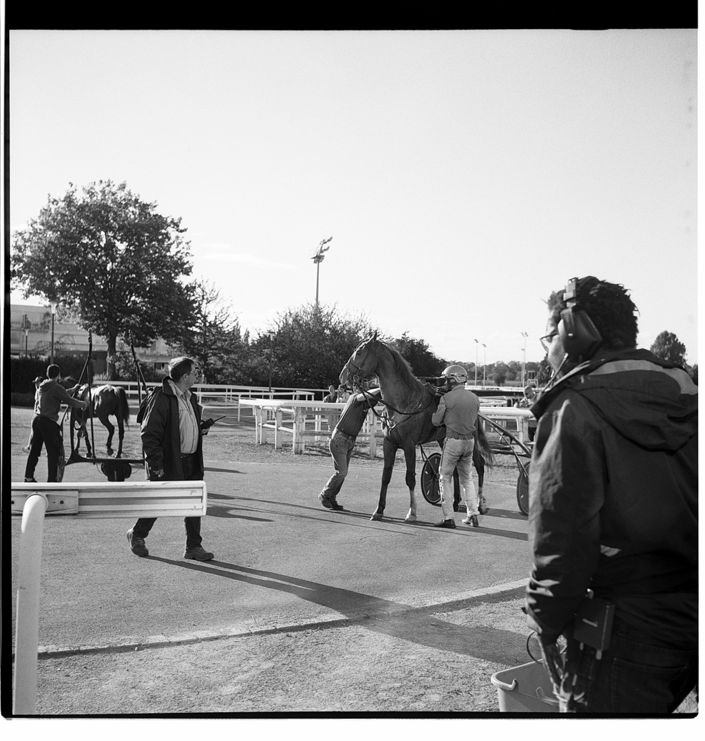 Photographie en noir et blanc d'un hippodrome, avec des hommes préparant des chevaux, un homme portant un équipement audio, et un environnement de course visible en arrière-plan.