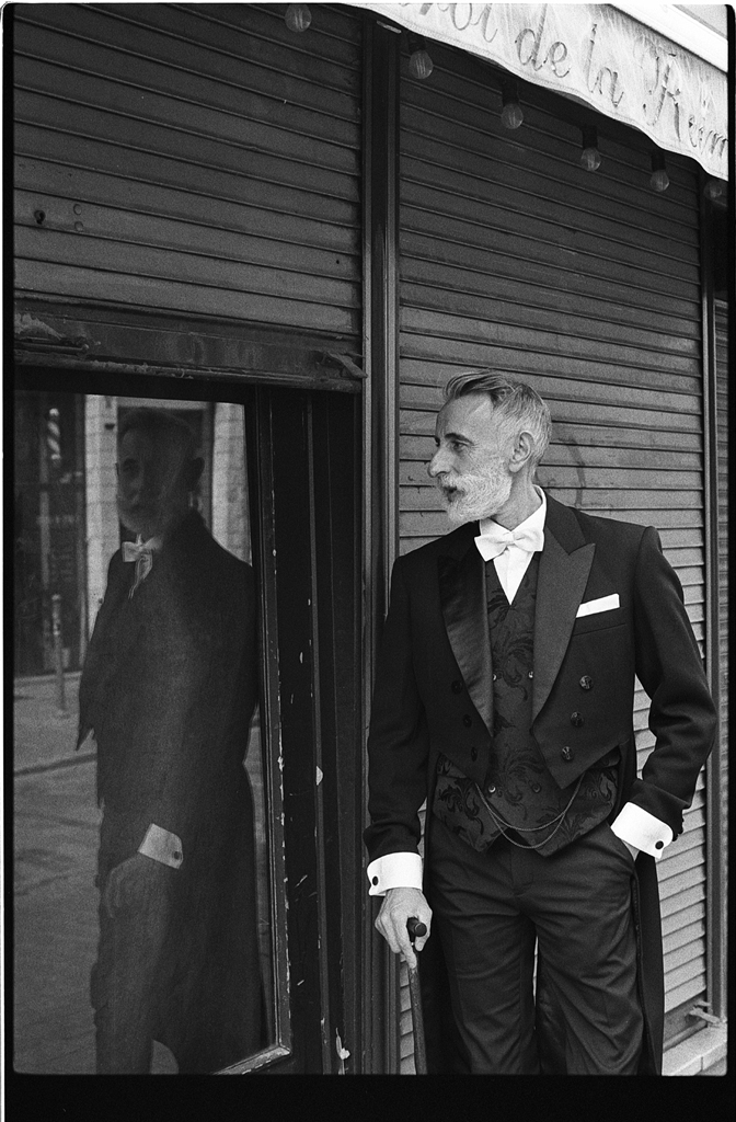 Un homme élégant en costume noir se tient à côté d'une vitrine, avec son reflet visible dans le verre. Il a une barbe grise et porte une cravate noire, semblant contempler l'environnement.