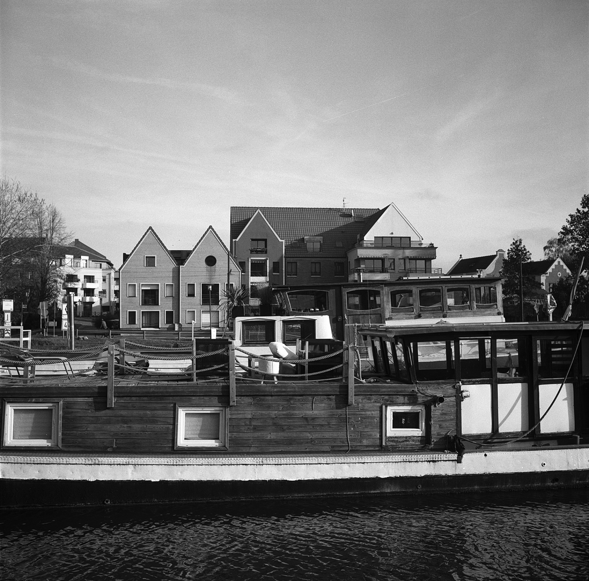 Un bateau en bois amarré sur un port, avec des bâtiments modernes en arrière-plan sous un ciel dégagé. Photo prise au Rolleiflex et pellicule Ilford FP4