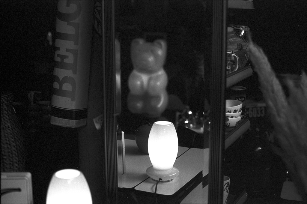 Photo en noir et blanc d'une lampe sur une table, avec un reflet d'un ours en peluche dans un miroir et divers objets en arrière-plan dans une boutique d'antiquités.