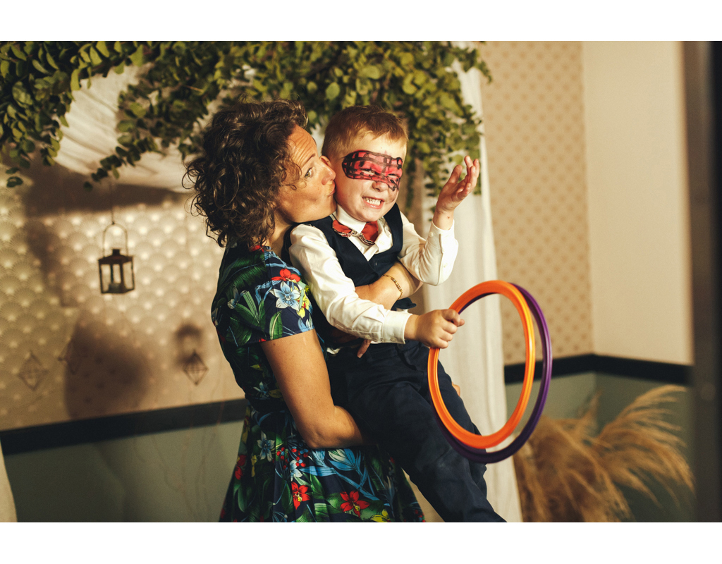 Une mère embrasse son fils, qui porte un masque, tout en tenant des anneaux colorés dans une pièce décorée avec des plantes.