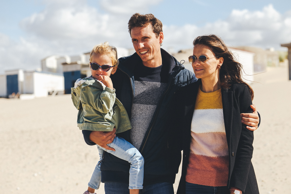 Une famille se tenant sur la plage, composée d'un homme, d'une femme et d'une petite fille dans les bras de l'homme. Ils sourient et portent des vêtements de saison, avec un ciel nuageux en arrière-plan.