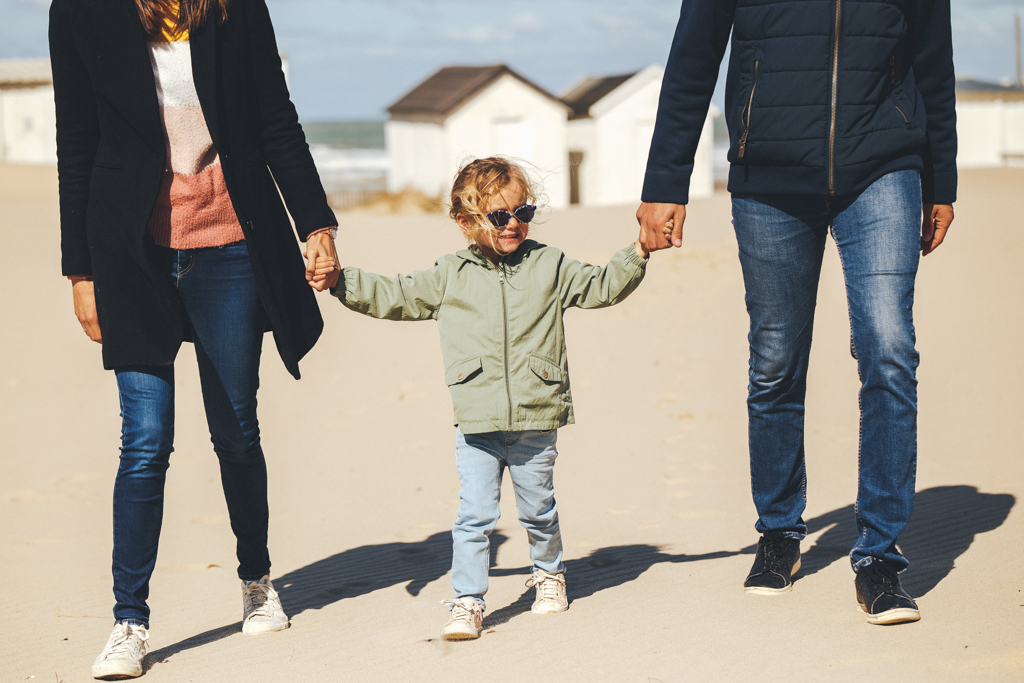 Une famille marchant main dans la main sur la plage, avec un enfant portant des lunettes de soleil et un manteau léger.