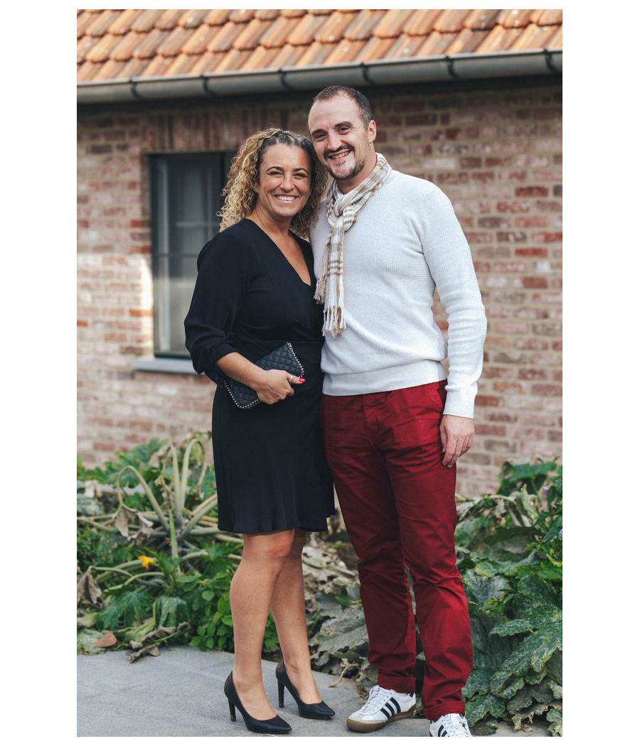 Un couple souriant pose ensemble en extérieur, la femme porte une robe noire et le homme un pull gris avec un pantalon rouge. Ils se tiennent l'un à côté de l'autre devant un bâtiment en briques.