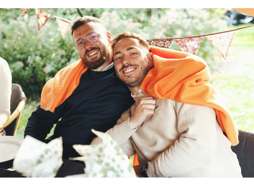 Deux hommes souriants posent ensemble, chacun portant une couverture orange sur les épaules, assis à une table à l'extérieur avec un décor de guirlande en arrière-plan.
