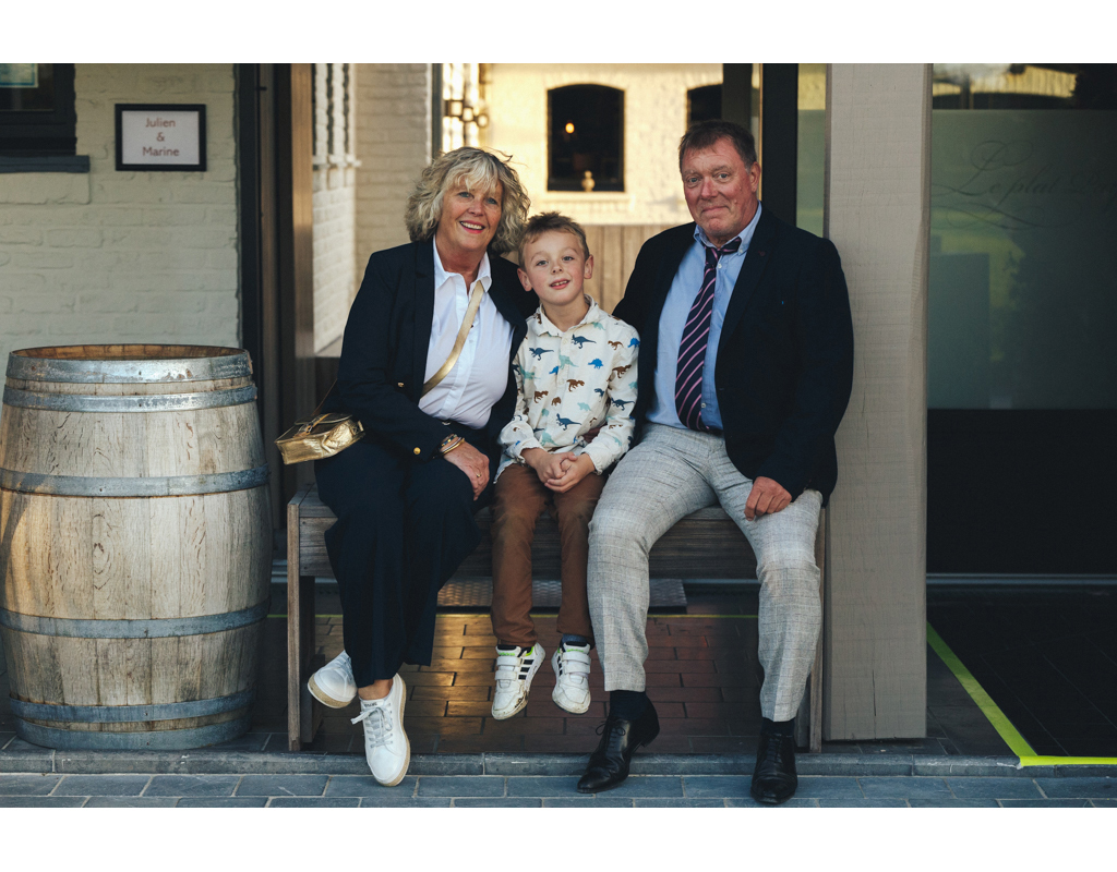 Un grand-parent, un enfant et un homme assis sur un banc à l'extérieur, souriant ensemble devant un bâtiment.