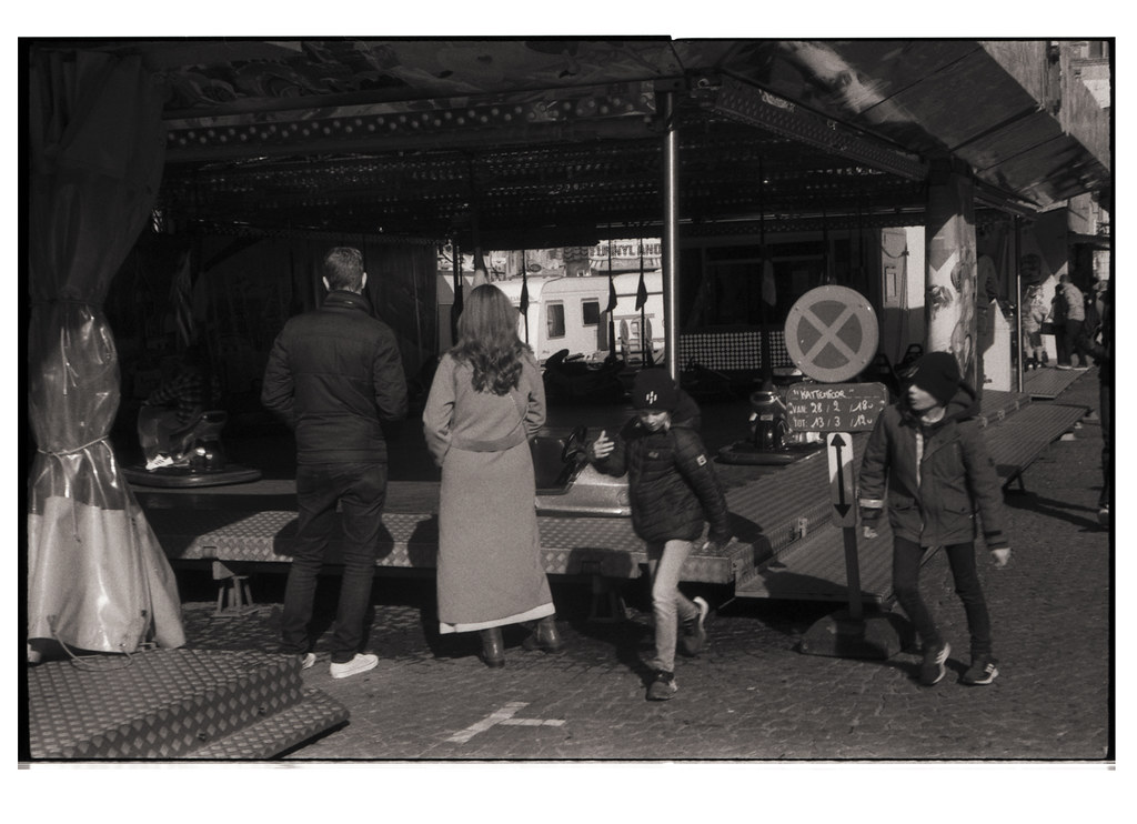 Une photographie en noir et blanc montrant des adultes et des enfants dans un environnement de fête foraine. On voit deux adultes de dos, un homme et une femme, ainsi que deux enfants en mouvement devant eux, près d'une attraction. Des éléments d'une fête foraine sont visibles en arrière-plan.