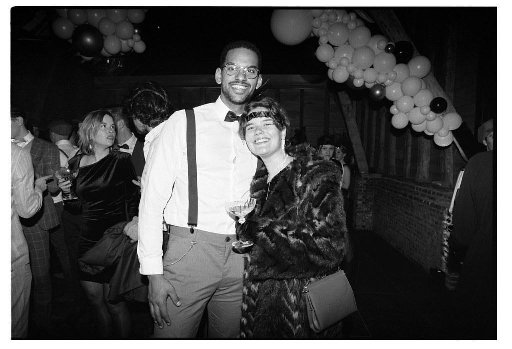 Un homme et une femme souriants tiennent des verres dans une ambiance de soirée festive, entourés de décorations et d'autres invités.