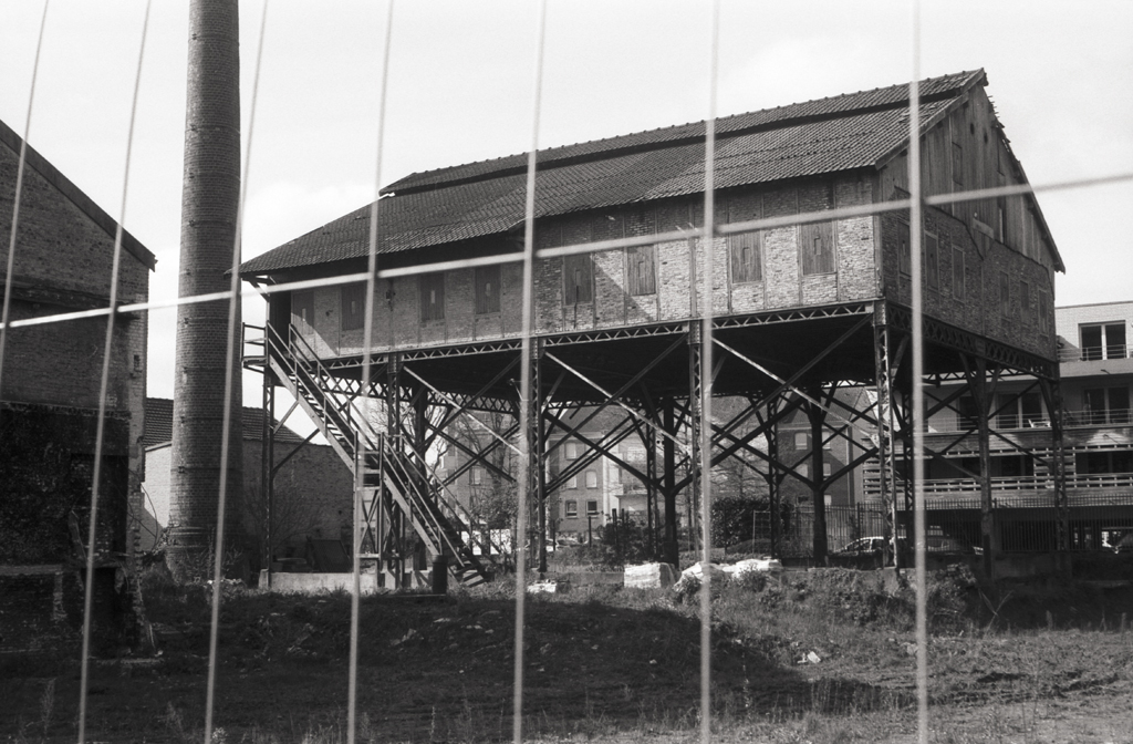 Un bâtiment sur pilotis avec une structure métallique, entouré d'une clôture, photographié en noir et blanc.