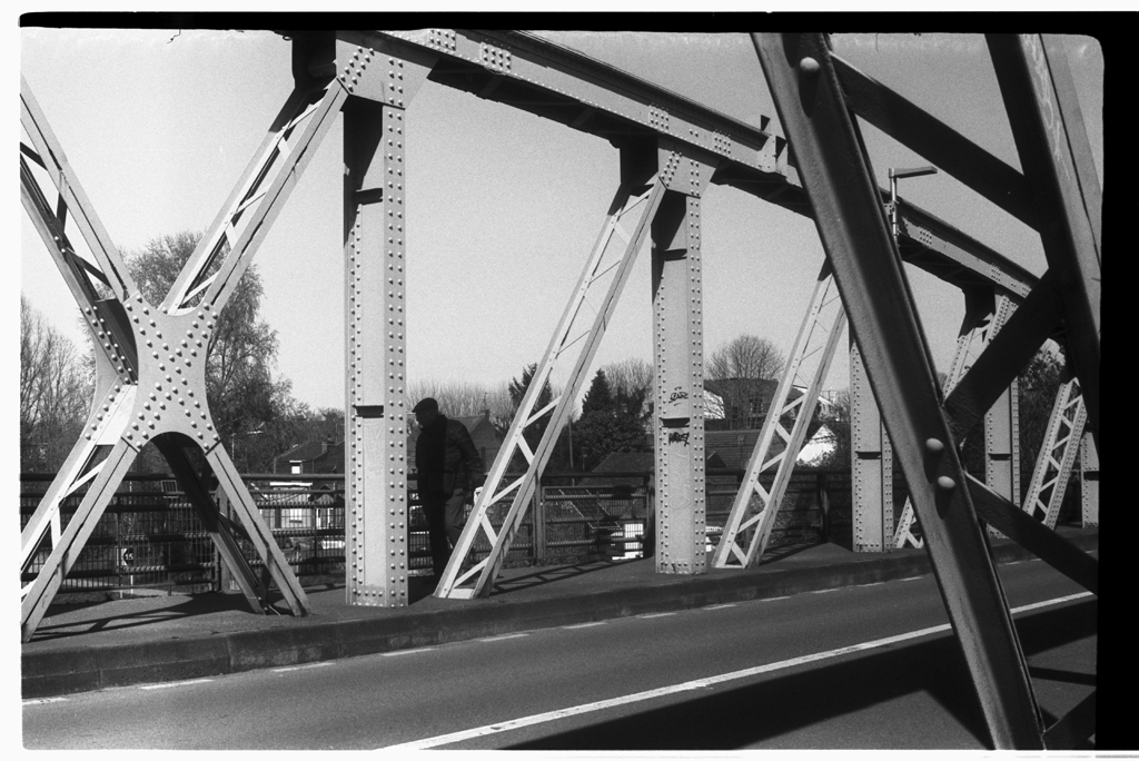 Un homme debout près des poutres en métal d'un pont, photographié en noir et blanc.