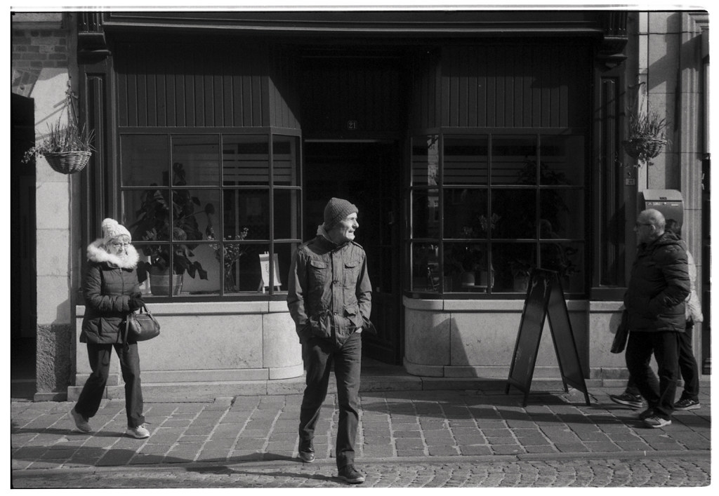 Une scène urbaine en noir et blanc montrant des passants marchant devant un café avec de grandes fenêtres. À gauche, une femme portant un manteau sobre et une écharpe, à droite, un homme âgé avec des lunettes. Au centre, un homme plus jeune, souriant et vêtu d'une veste en denim. En arrière-plan, la façade d'un café avec une enseigne sur le trottoir.
