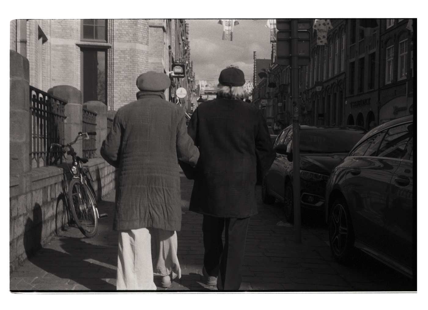 Deux personnes âgées marchant côte à côte dans une rue, portant des manteaux et des chapeaux, capturées en noir et blanc.