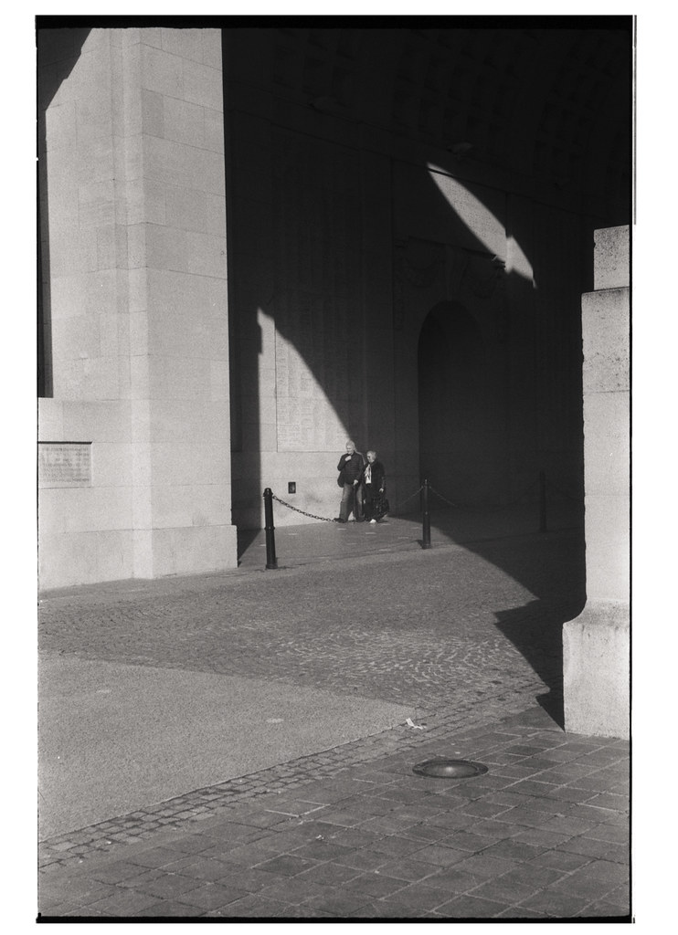 Deux personnes se tenant près d'une structure en pierre, avec de longues ombres projetées sur le sol en pavés.