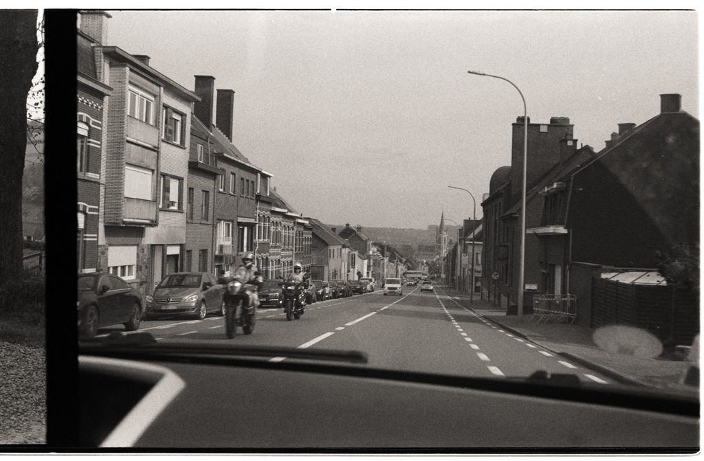 Vue d'une route à travers la vitre d'une voiture, avec des maisons et des véhicules sur les côtés. Impression en noir et blanc.