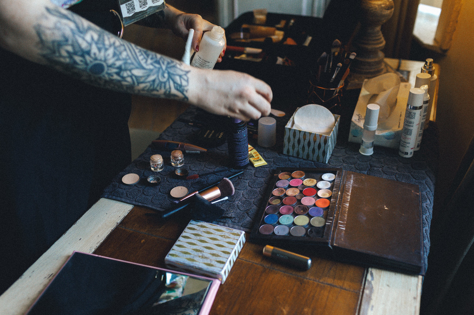 Un close-up d'une main appliquant du maquillage sur une table chargée de produits de beauté, avec divers pinceaux et palettes de maquillage en arrière-plan.