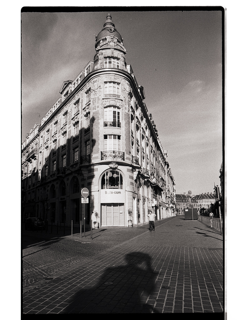 Une photographie en noir et blanc d'un bâtiment historique aux angles arrondis, capturée sur une rue pavée, avec une ombre projetée au premier plan.