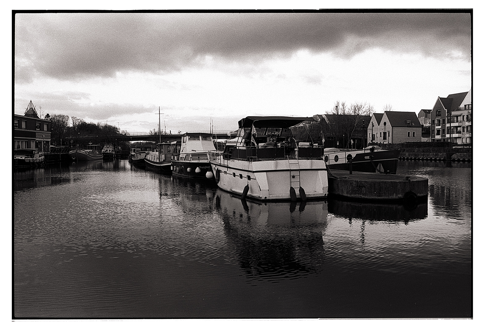 Scène en noir et blanc montrant des bateaux amarrés le long d'un canal, avec des bâtiments en arrière-plan sous un ciel nuageux.