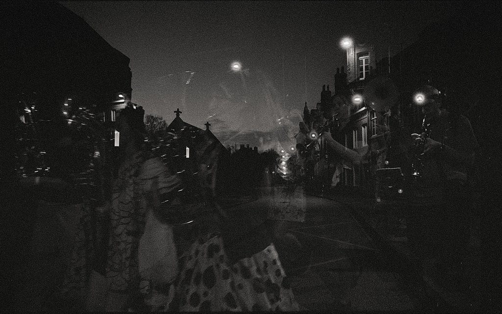 Photographie en noir et blanc illustrant une scène urbaine avec des superpositions abstraites, montrant des silhouettes de personnes et de bâtiments au crépuscule.