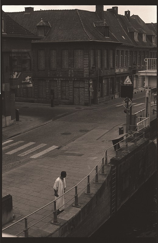 Une rue urbaine avec des bâtiments historiques en arrière-plan, un piéton vêtu d'une robe blanche marchant le long d'un mur près d'un canal.